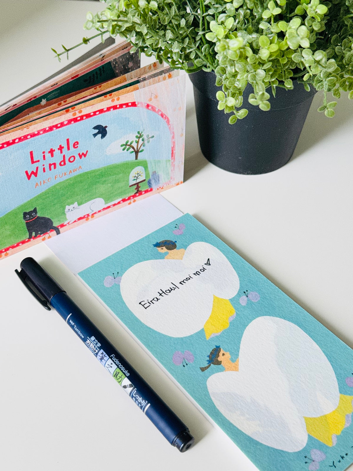 A styled flat-lay featuring a children's book titled 'Little Window' by Jill Redpath, a light blue notepad with duck illustrations and the text 'Eve-Mae now she's a duck', a black pen, and a potted green plant on a white surface.