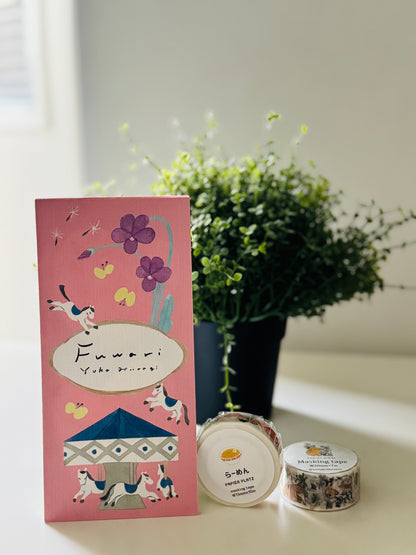 A pink illustrated card or package standing on a white surface beside a potted green plant, with two small round jars in front. The package has whimsical drawings of flowers, birds, and a carousel, and the visible text includes 'Fuwemu Yoko Mugo!' and 'Sakura'.