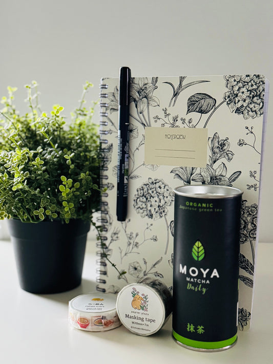 A styled flat-lay of a desk setup featuring a black metal tin of Moya Matcha Green Tea, a floral-patterned notepad, a black pen, a small tin of masking tape, and a potted green plant on a white surface against a light gray background.