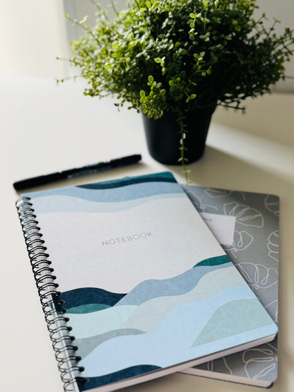 A spiral-bound notebook with a blue and teal wavy mountain pattern on the cover, placed on a light-colored desk next to a black pen and a small potted green plant.