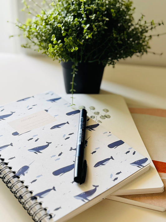 A spiral-bound notebook with a white cover decorated with a navy blue whale pattern, resting on a desk next to a black pen. A small potted green plant is visible in the background.