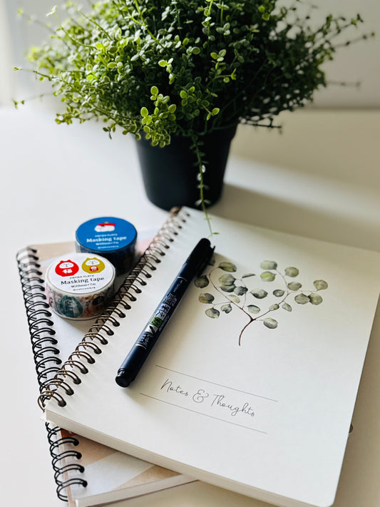 A styled flat-lay of a white spiral notebook with a watercolor eucalyptus branch illustration and the words 'Notes & Thoughts' written on the cover. A black pen rests on the notebook, alongside two rolls of washi tape, with a potted green plant in the background on a white surface.