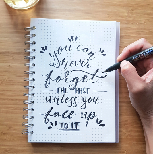 Eira Haul Tombow Fudenosuke soft tip brush pen creating hand lettering in a dot grid notebook on a wooden desk