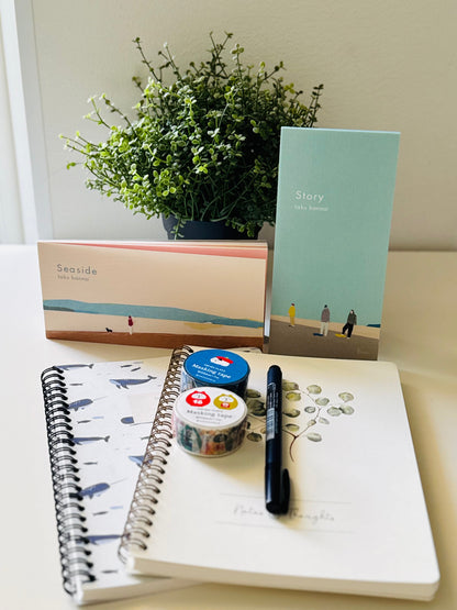 A styled stationery and gift set arranged on a tabletop in front of a potted plant. The set includes a notebook with a blue whale pattern, a mint green card or notebook titled 'Story', a peach and blue box labeled 'Seaside', a white notebook with handwritten notes, a black pen, and two small decorative washi tape rolls.