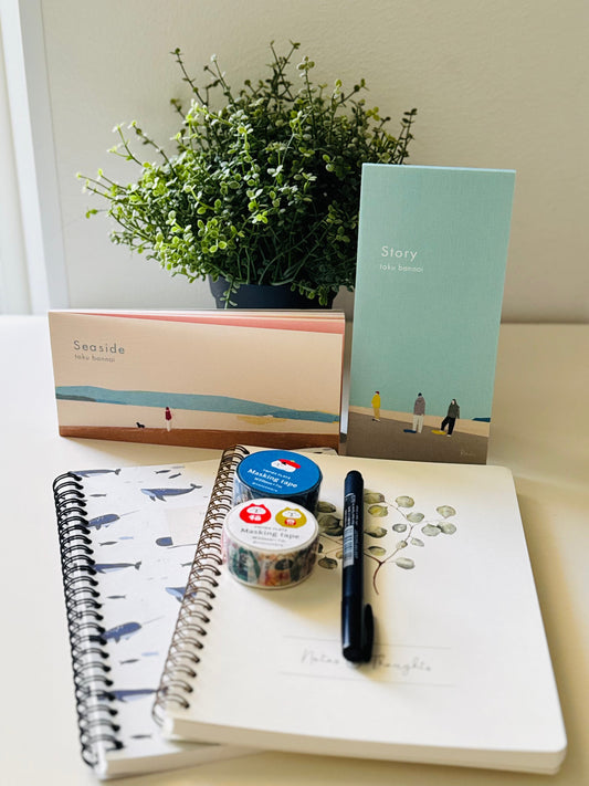 A styled stationery and gift set arranged on a tabletop in front of a potted plant. The set includes a notebook with a blue whale pattern, a mint green card or notebook titled 'Story', a peach and blue box labeled 'Seaside', a white notebook with handwritten notes, a black pen, and two small decorative washi tape rolls.