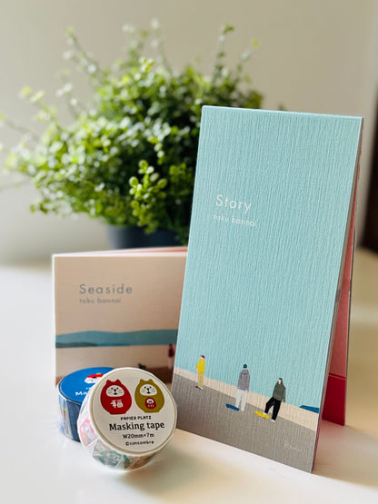 A styled flat-lay of Seaside Take Dancer stationery items on a light surface with a blurred green plant in the background. The set includes a light blue notebook with a beach illustration and the title 'Story', a cream-colored box labeled 'Seaside', and a roll of washi tape with cat illustrations and the text 'masking tape'.