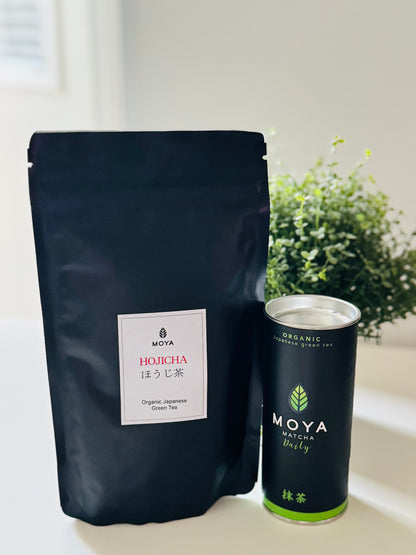 A black stand-up pouch of Moya Hojicha Japanese green tea next to a matching black canister of Moya Matcha Daily, set on a white surface with a blurred green plant in the background.