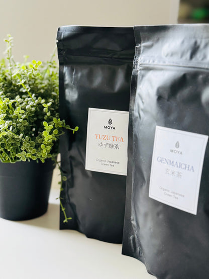 Two black stand-up pouches of Moya tea, one labeled Yuzu Tea and the other Genmaicha, standing on a white surface next to a potted green plant.