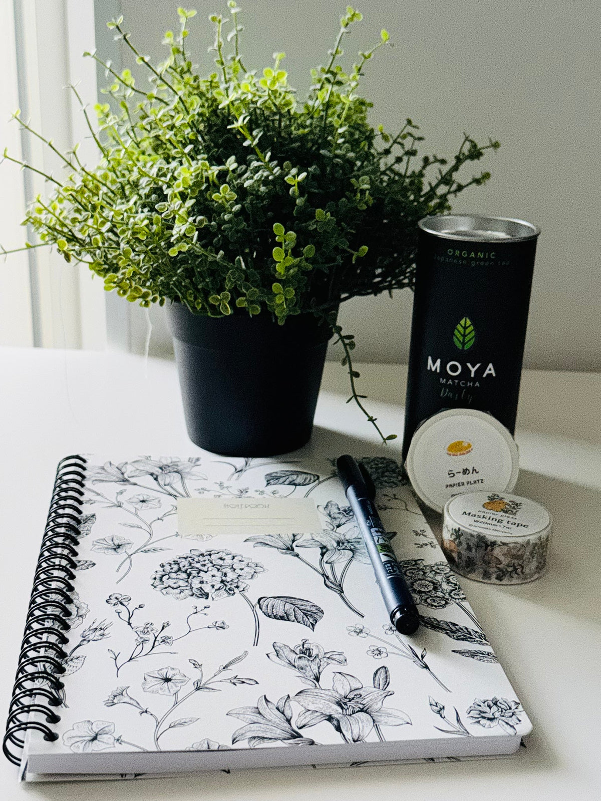 A styled flat-lay of a floral spiral notebook with a black pen, a black tin of Moya Naturals Organic Green Tea, a small jar of S-BA Premium Supplements, and a potted green plant on a white surface.